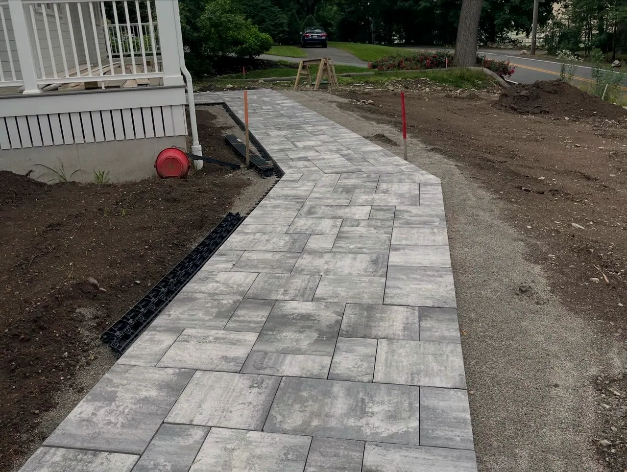 Gray paver walkway being laid along a porch, edge restraint and layout stakes in place