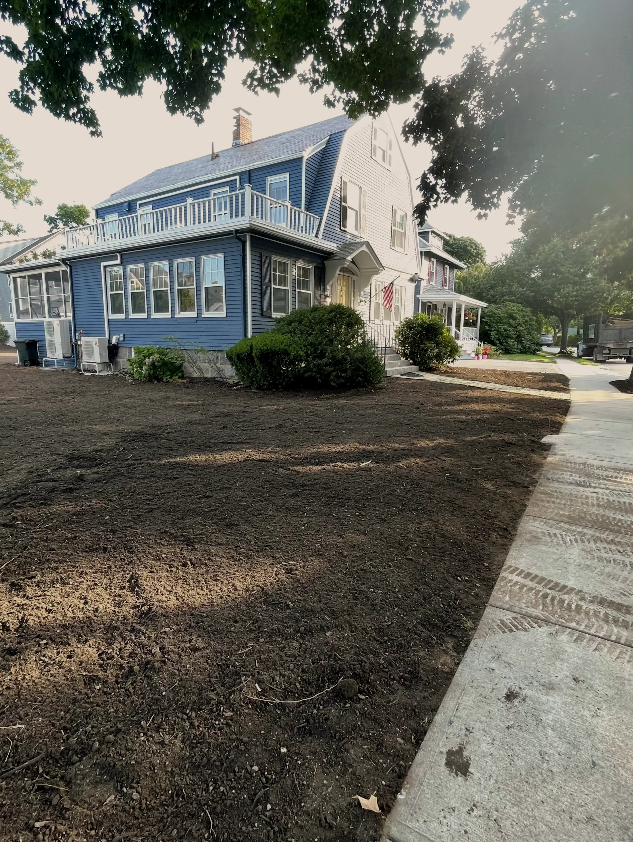 Side yard graded to bare loam next to a blue house and concrete sidewalk